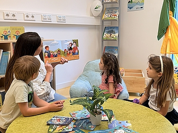 Sozialpädagogische Assistenzkraft Fazile Avdulli liest drei Kindern im Fröbel-Kindergarten Am Kalkbruche aus dem Buch „Das Farbmonster“ vor. Sie sitzen gemeinsam an einem runden grünen Tisch in der Kinderbibliothek.